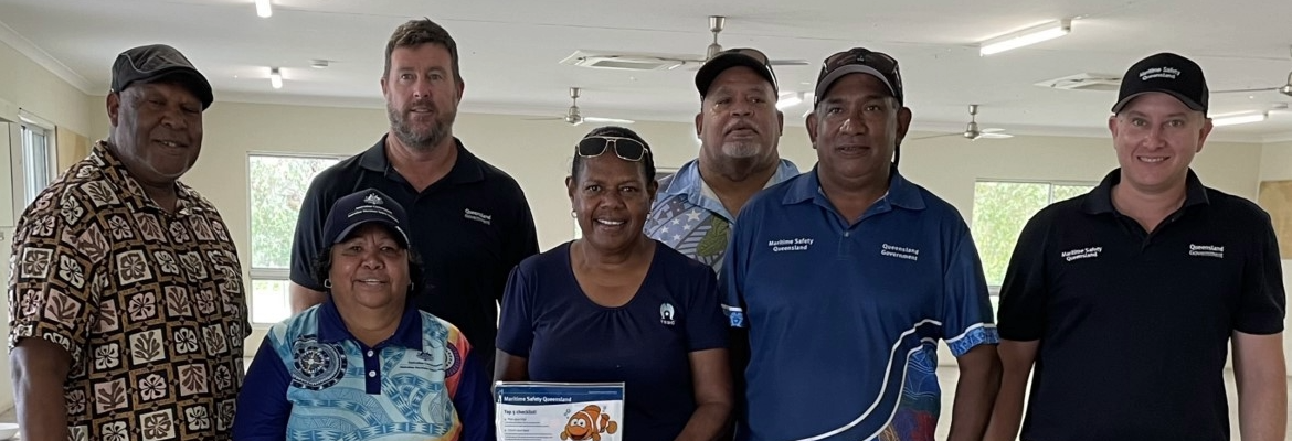 A group of seven people stand indoors, smiling at the camera. Some wear uniform with logos. One person in the center holds a certificate with a fish illustration. The background has white walls, windows, and ceiling fans.